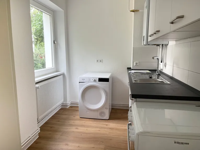Blick in einen Raum, links ein Fenster, hinten eine Waschmaschine, rechts eine K&uuml;chzeile.