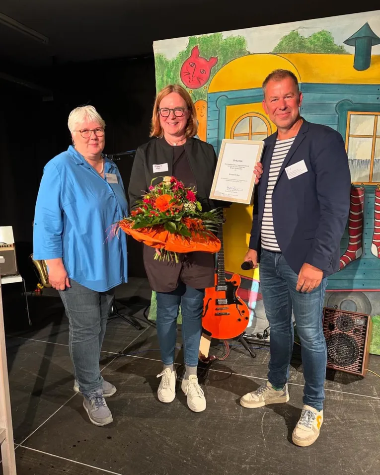 Drei Menschen nebeneinander. Die Frau in der Mitte hat eine Urkunde und einen Blumenstrau&szlig; &uuml;berreicht bekommen.