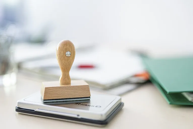 Im Vordergrund im Fokus ein klassische Stempel aus Holz. Er steht auf einem Stempelkissen. Im Hintergrund unscharf Aktenordner.