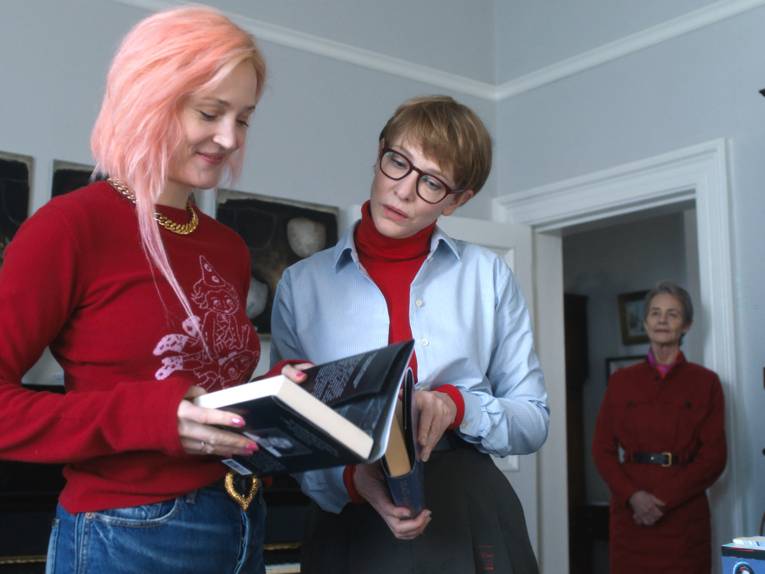 Eine junge Frau guckt auf die erste Seite eines Buches und lächelt. Eine andere Frau guckt auch in das Buch. Eine ältere Frau steht abseits und guckt den beiden zu.