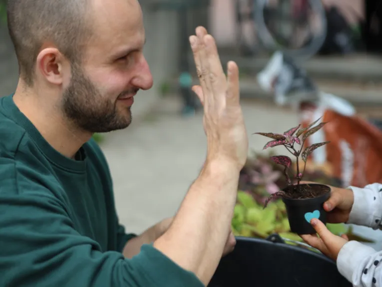 Ein Mann von der Seite, er h&auml;lt eine Hand hoch und zeigt f&uuml;nf Finger. Davor ein paar Kinderh&auml;nde, die einem Blumentopf mit einer Pflanze halten.
