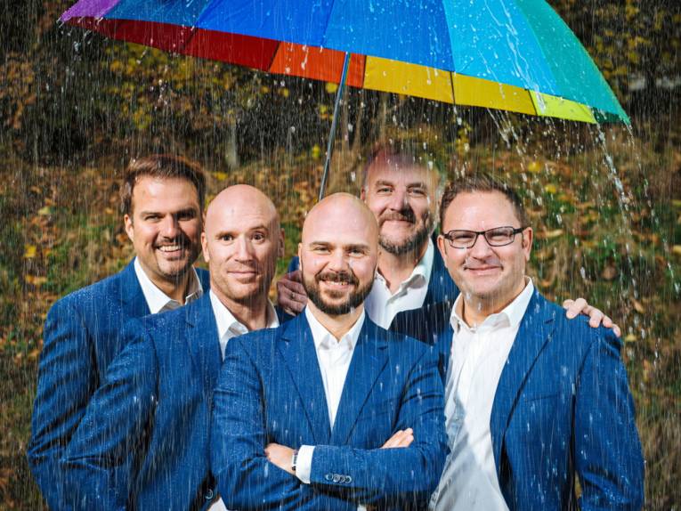 Fünf Männer unterschiedlichen Alters stehen in blauen Anzügen unter einem bunten Schirm im Regen.