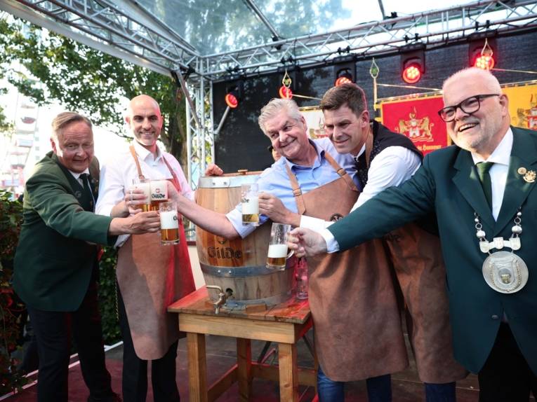 Fünf Männer an einem Bierfass. Sie schwenken die Gläser.