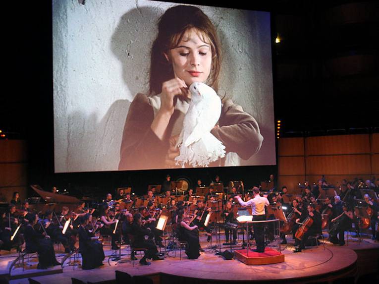 Ausschnitt aus dem Film Drei "Haselnüsse für Aschenbrödel" auf einer großen Leiinwand, darunter ein Sinfonieorchester.