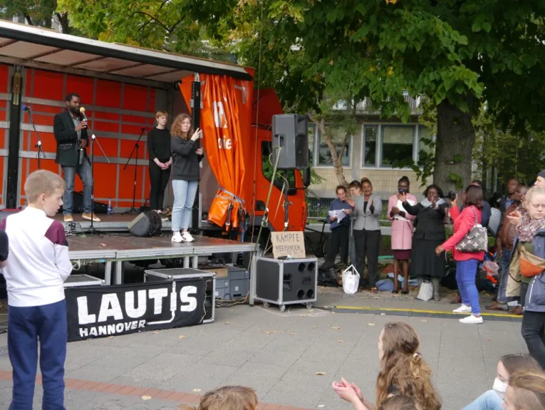 Eine Person auf einer B&uuml;hne spricht vor eine Gruppe Personen