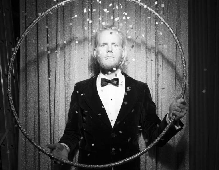 Mann mit langem Haar und Bart in Hemd, Fliege und Jackett hält einen großen Reifen.