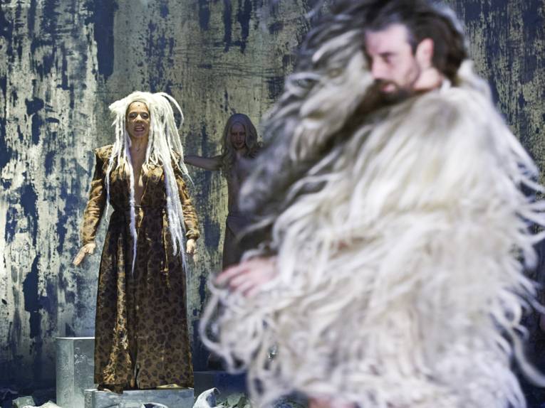 Eine Frau mit langem Haar auf einer Bühne, im Vordergrund unscharf ein Mann in einem Fellkostüm