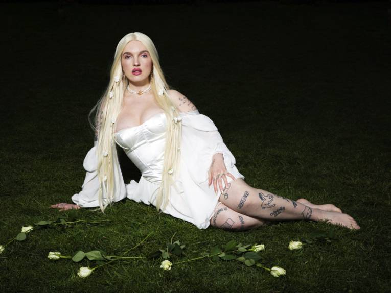 Blonde Frau in weißem Kleid mit vielen Tattoos liegt auf dem Rasen.
