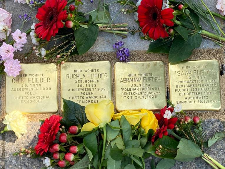 Vier goldene, quadratische Steine mit Inschrift im Boden: Chin Flieder, Ruchla Flieder, Abraham Flieder und Isaak Flieder.  Blumen drum herum.