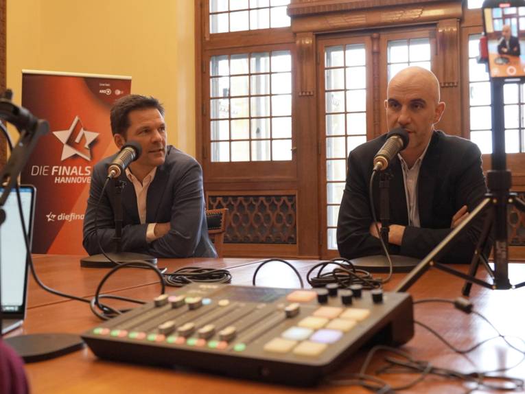 Zwei Männer sitzen an einem Tisch, vor ihnen Mikrofone und ein technisches flaches Bord mit Schaltern und Reglern. Ein Mann spricht, der andere sieht ihn an.