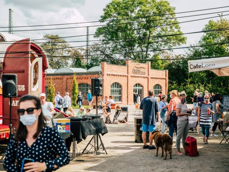 Personen auf einem Außengelände an Verkaufsständen, teilweise mit Masken.
