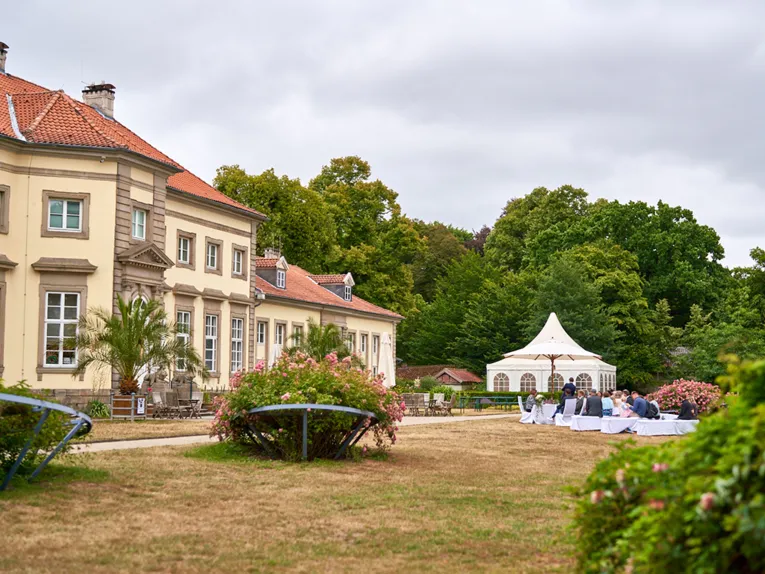 Ein mit Blumen und Kerzen geschm&uuml;ckter Tisch und drei St&uuml;hle mit wei&szlig;en Stuhlhussen im Gr&uuml;nen.