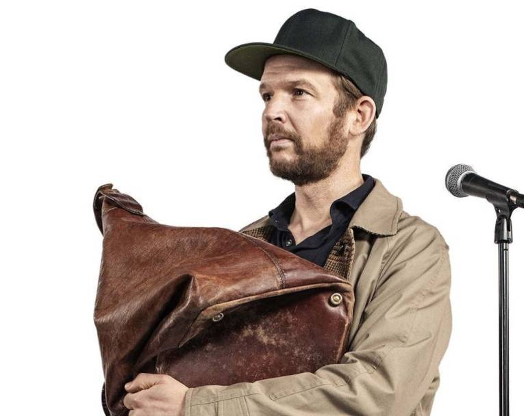 Mann mit Vollbart und Cap hält eine alte Ledertasche in den Armen, neben ihm steht ein Mikro.