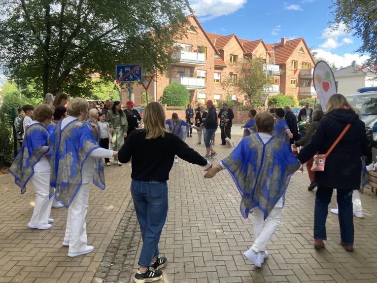 Im Kreis tanzen die Damen mit den Besuchenden. Die Damne tragen wei&szlig;e Kleidung mit einem blauen seidenden Poncho mit Mustern.