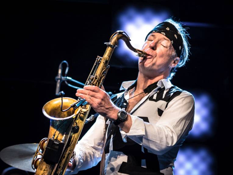 Mann mit Stirnband und geschlossenen Augen spielt Saxophon.