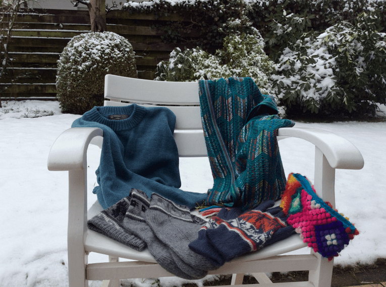 Strickpulli, Handschuhe, Socken, Schal und Strinband auf einem Stuhl in winterlicher Umgebung.