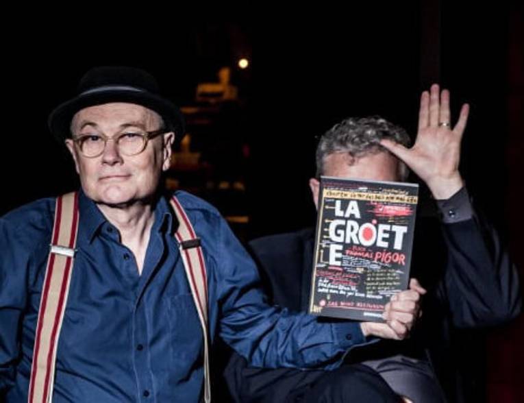 Ein Mann mit Brille, Hut, Hemd und Hosenträgern hält ein Heft mit der Schrift La Groete vor das Gesicht eines zweiten mit kurzem Haar, Hemd und Jackett.