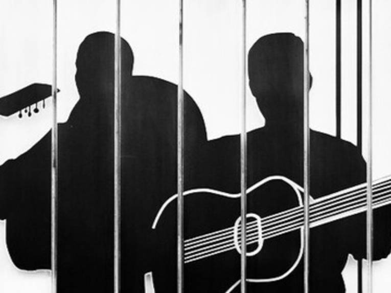 Schwarze Silhouette von zwei Männern hinter senkrechten Stäben, einer hält eine Gitarre.