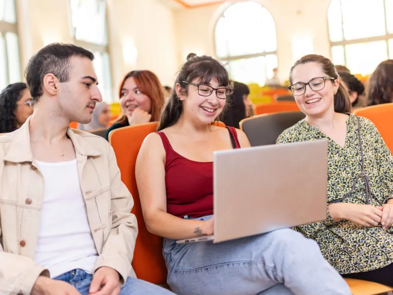 Zwei Frauen und ein Mann sitzen bei einer Veranstaltung in der vorderen Reihe und schauen gemeinsam auf einen Laptop.