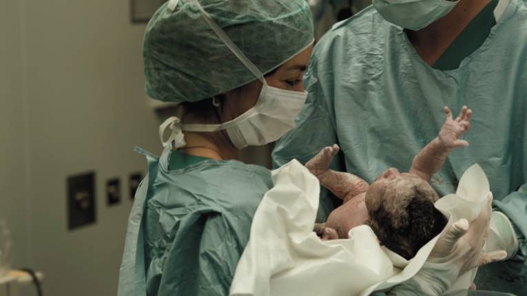 Frau in medizinischer Schutzkleidung hält ein Neugeborenes unmittelbar nach der Geburt in den Händen.