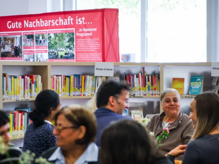 Vor einem B&uuml;cherregal sitzen Menschen, die l&auml;cheln und miteinander sprechen. Im Hintergrund ein rotes Transparent "Gute Nachbarschaft ist ...".