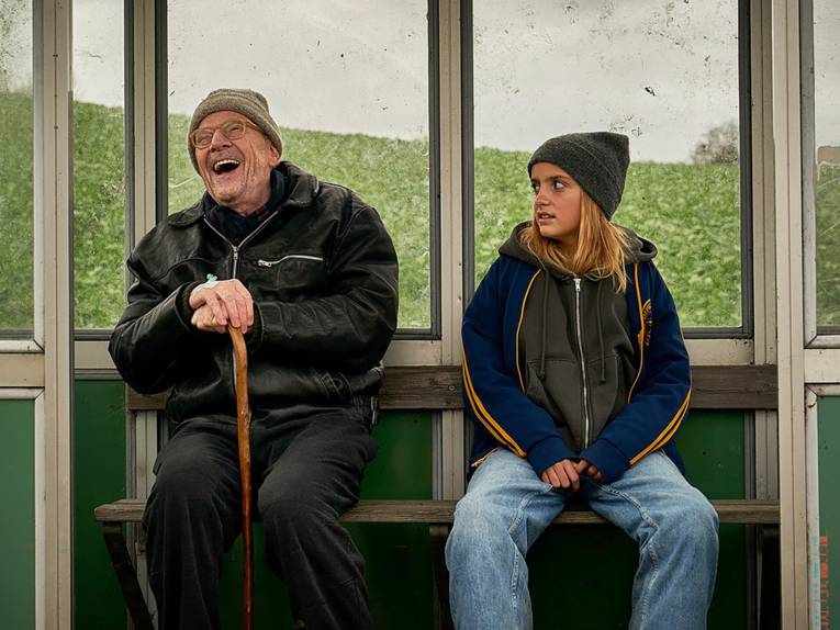 Ein Mann und ein Mädchen sitzen in einem Wartehäuschen. Der Mann lacht schallend, das Mädchen guckt ihn skeptisch an.