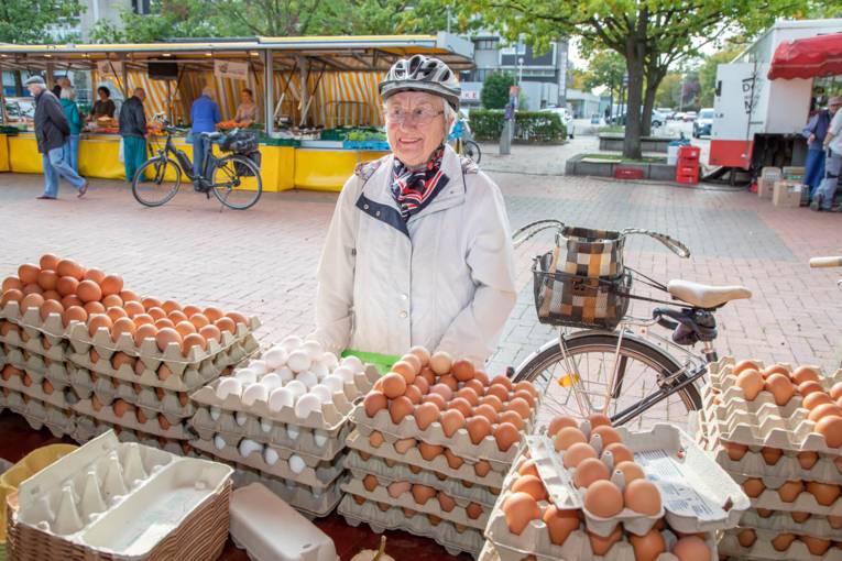 Eine Rentnerin mit Fahrradhelm auf dem Kopf steht neben ihrem Rad und vor einem Marktstand mit Eiern und lacht in die Kamera.