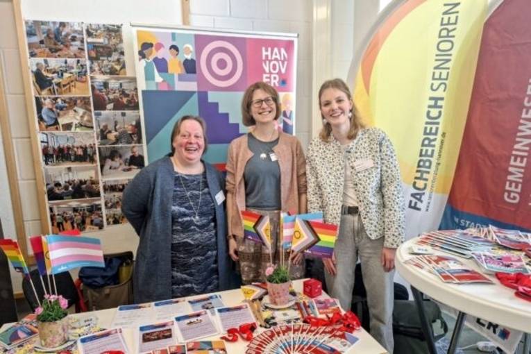 Drei Frauen stehen vor Bildern und Werbeflächen des KSH. Im Vordergrund sind zwei Tische mit verschiedenen Broschüren und Flyern sowie Queer-freundlichen Fähnchen.