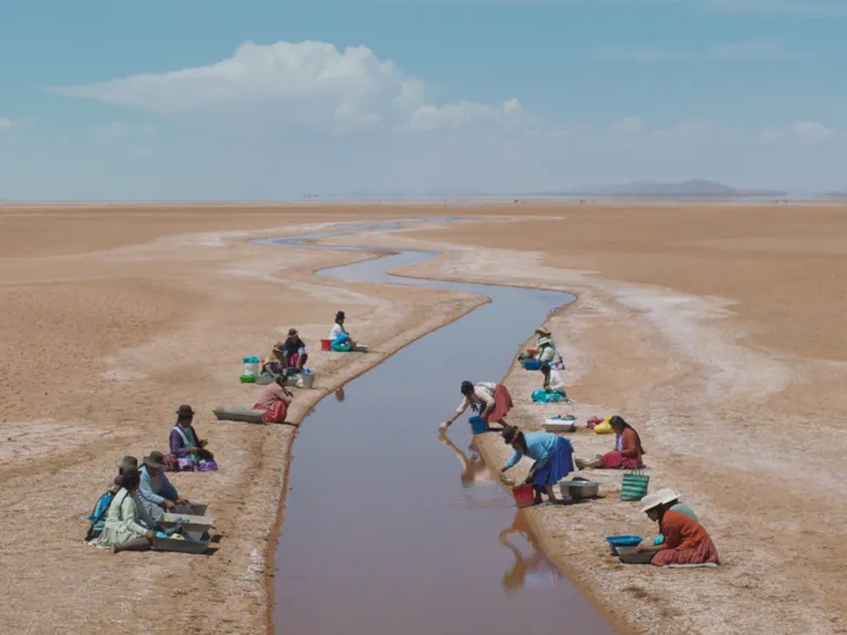 Menschen sitzen entlang eines schmalen Wasserlaufs in einer weiten, trockenen Ebene und arbeiten mit Schalen am Ufer. Die karge Landschaft, der sich durch das Bild schlängelnde Kanal und der weite Himmel erzeugen eine eindrucksvolle Stimmung von Weite und Verbundenheit mit der Natur.