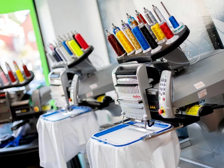 Stoff ist in einen Rahmen gespannt, eine Maschine bestickt den gespannten Stoff innerhalb des Rahmens. Auf der Maschine sind verschiedenfarbige Garnrollen.