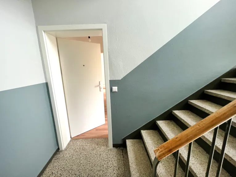 Ein Treppenhaus mit einem grau-blauem Streifen an der Wand, die T&uuml;r zu einer Wohnung ist ge&ouml;ffnet.
