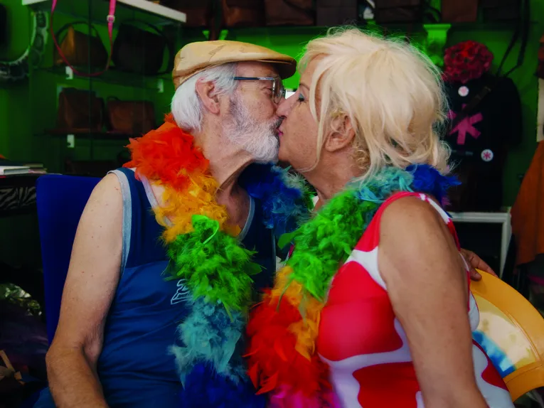 Das Bild zeigt zwei ältere Personen, die sich in einem Innenraum küssen. Beide tragen bunte Federboas in den Farben des Regenbogens. Eine Person trägt eine helle Schiebermütze und eine Brille, die andere ein rot-weiß gemustertes Kleidungsstück. Im Hintergrund sind Regale mit Taschen und weitere dekorative Elemente zu erkennen. Die farbenfrohe Szene wird durch warmes Licht und kräftige Farben geprägt.
