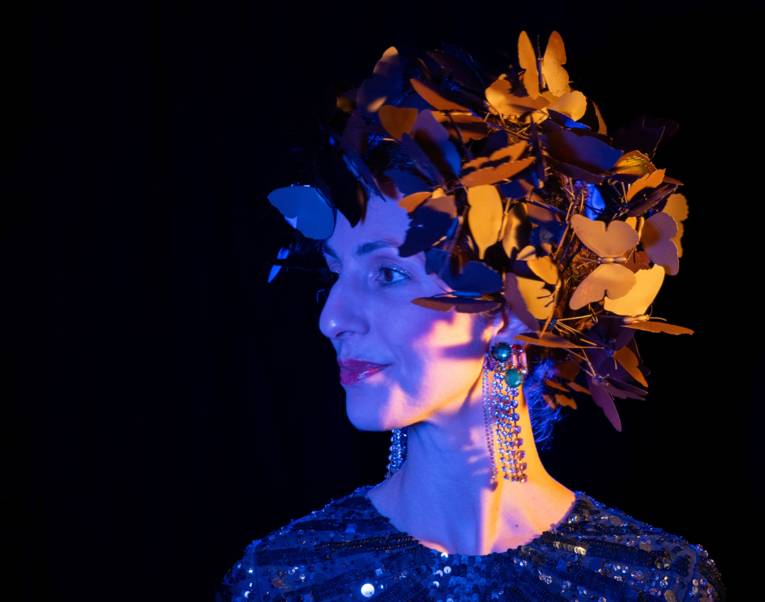 Leicht seitliches Porträt einer Frau mit Kopfschmuck aus blätterartigen Elementen und langen Ohrringen.