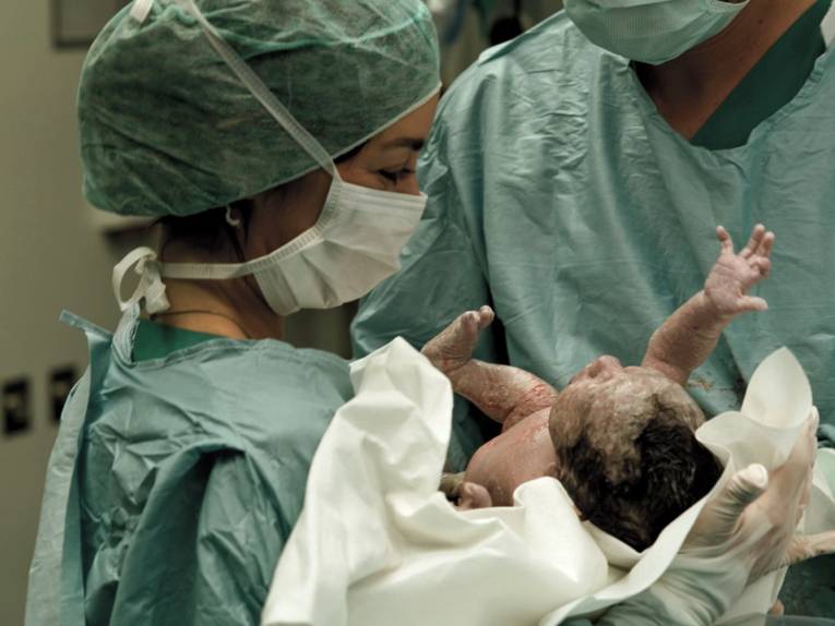 Frau in medizinischer Schutzkleidung hält ein Neugeborenes unmittelbar nach der Geburt in den Händen.