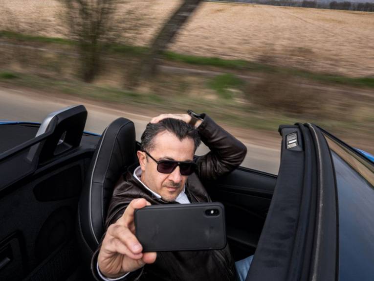 Ein Mann fährt in einem Sportwagen und macht dabei ein Selfie von sich.