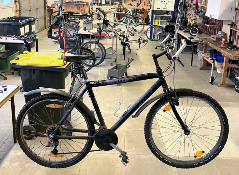 Ein Herrenrad hängt an Sattel und Lenker an zwei Ketten und schwebt leicht über dem Boden einer Werkstatt, dahinter sind weitere Räder, Werkbänke, Mülltonnen und Werkzeuge zu sehen.