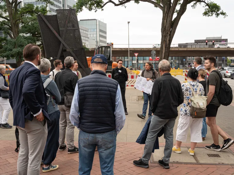 Eine Gruppe von Menschen steht auf einem B&uuml;rgersteig. Alle blicken auf einen Mann in der Mitte, der eine Karte h&auml;lt. Hinter ihm verl&auml;uft eine Hochstra&szlig;e, rechts eine Stra&szlig;e, auf beiden fahren Autos.