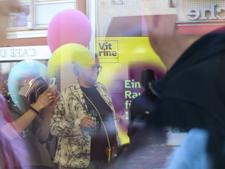 Mehrere Menschen, einige Luftballons und ein Aufsteller. Fotografiert durch eine Glasscheibe. Formen und Farben verschwimmen leicht und ergeben eine Art Collage. Die Farben gelb und lila dominieren das Bild.