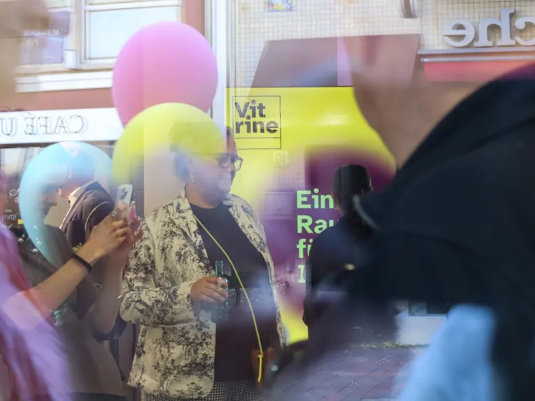Mehrere Menschen, einige Luftballons und ein Aufsteller. Fotografiert durch eine Glasscheibe. Formen und Farben verschwimmen leicht und ergeben eine Art Collage. Die Farben gelb und lila dominieren das Bild.