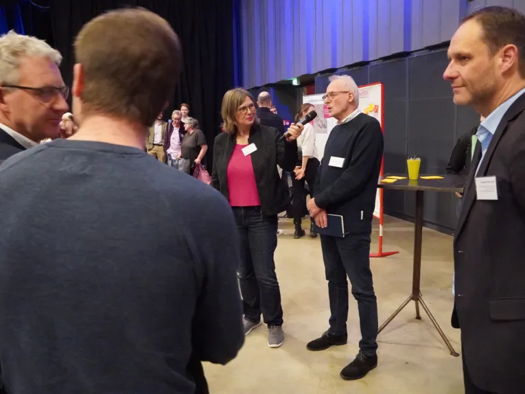 Eine Frau h&auml;lt einem Mann ein Mikrofon vor, Menschen stehen drum herum.