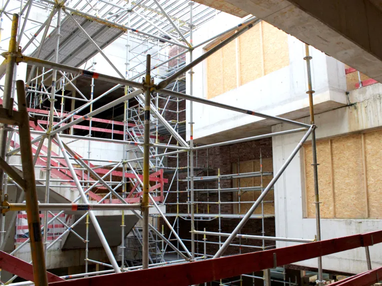 Zwei offene Geschosse in einem Rohbau mit einer Treppe links und mit Pressholz verschlossenen Fenstern rechts, die Mitte f&uuml;llt ein Bauger&uuml;st, von oben f&auml;llt Licht durch eine Metallkonstruktion.