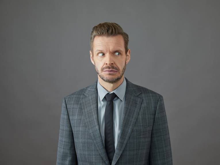 Zu sehen ist ein mittelalter Mann im Anzug vor einer grauen Wand.