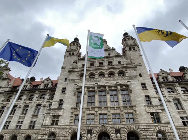 Die gr&uuml;n-wei&szlig;e-Mayors-for-Peace-Flagge weht gemeinsam mit einer Europa-Flagge, einer Ukraine-Flagge sowie der blau-gelben Stadtflagge der Stadt Leipzig vor einem prunkvollen Geb&auml;ude in Leipzig im Wind.