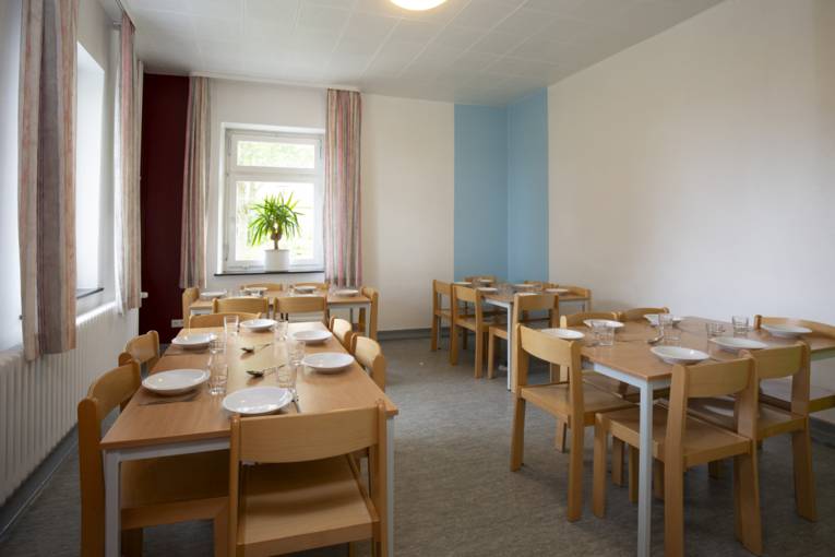 Gedeckter Speiseraum mit 4 Tischen und an den Tischen 5 - 6 Stühle. Auf den Tischen steht Geschirr.