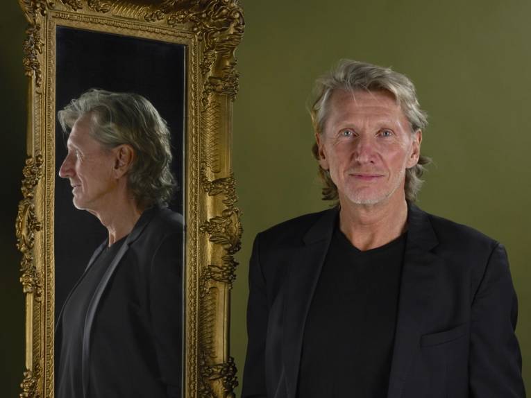 Mann mit graumeliertem Haar neben einem opulent gerahmten Spiegel mit seiner Profilansicht.