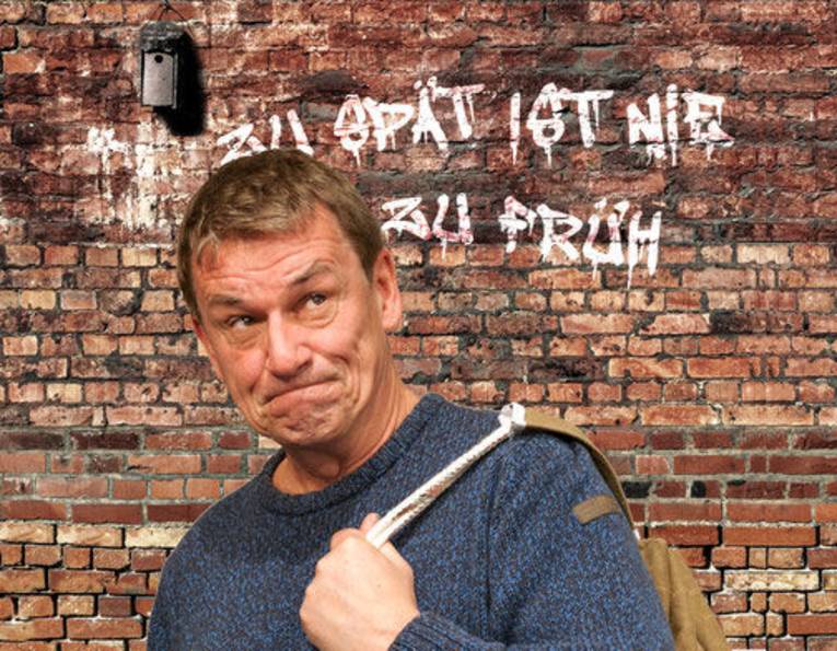 Grimassierender Mann mit kurzem Haar und Rucksack über der Schulter vor einer Backsteinwand mit Graffito Zu spät ist nie zu früh.