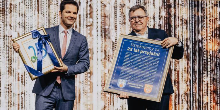 Zwei Personen auf einer Bühne, beide halten eine Art Auszeichnung in den Händen