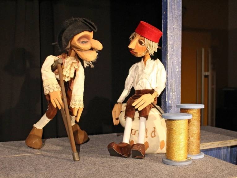 Zu sehen sind zwei Marionetten-Puppen: Eine Figur, die auf Krücken steht und ein Jüngling.