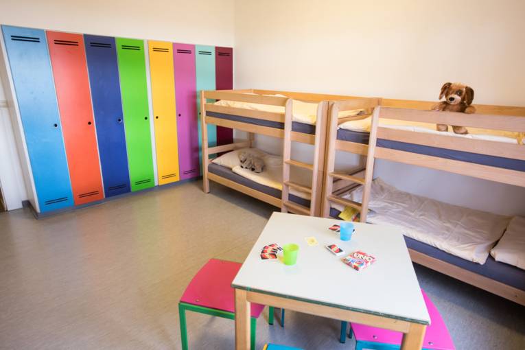 Mehrbettzimmer mit zwei Stockbetten und bunten Schränken. Im Vordergrund ein Tisch mit Hockern und einem Spiel