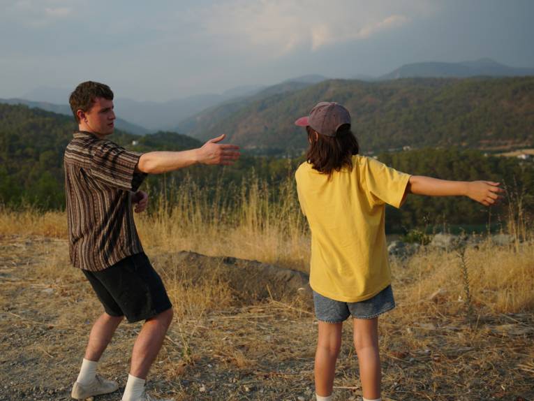 Zwei Menschen stehen auf einem trockenen Hügel mit Blick auf eine weite Berglandschaft. Mit ausgestreckten Armen vor der beeindruckenden Naturkulisse wirkt der Moment wie eine spielerische Begegnung mit der Freiheit. Warmes Abendlicht und die sanften Farben der Landschaft verleihen dem Bild eine ruhige, atmosphärische Stimmung.